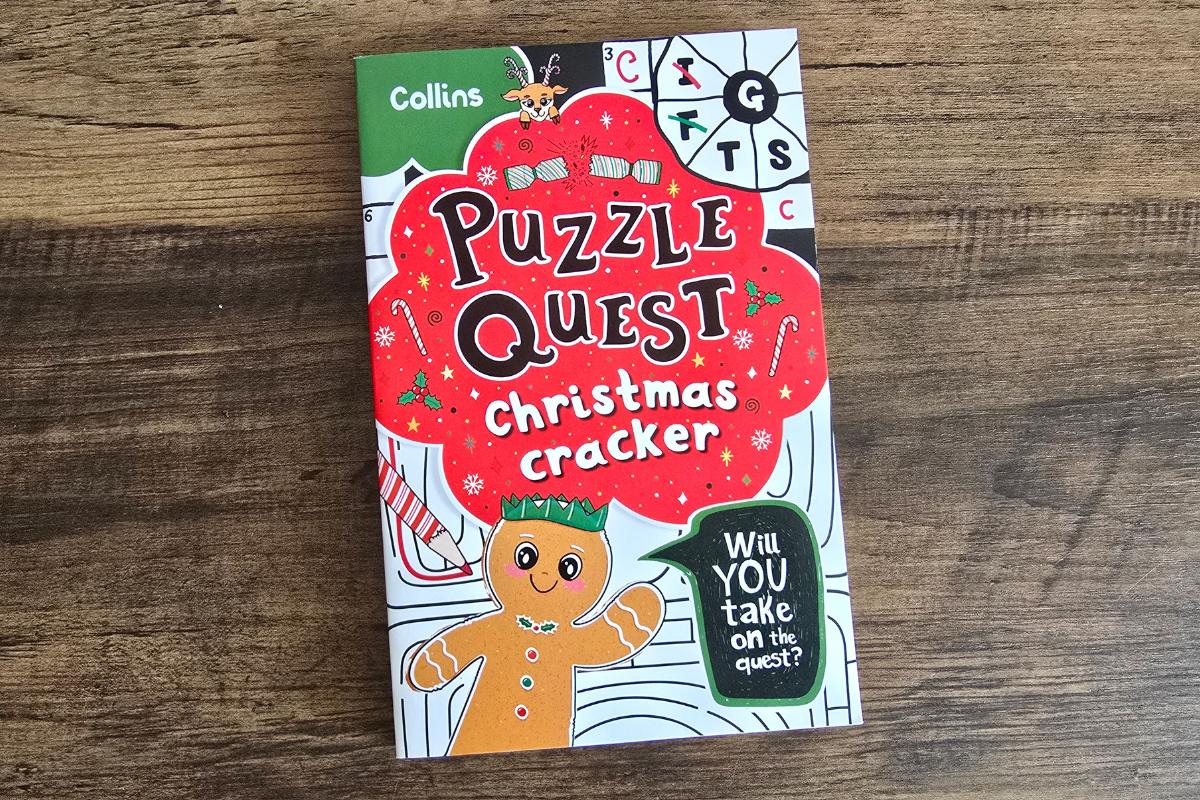 A colorful book titled "Puzzle Quest Christmas Cracker" by Collins lies on a wooden surface. Perfect for children's Christmas gifts, the cover features a smiling gingerbread figure and festive decorations with the tagline "Will YOU take on the quest?.