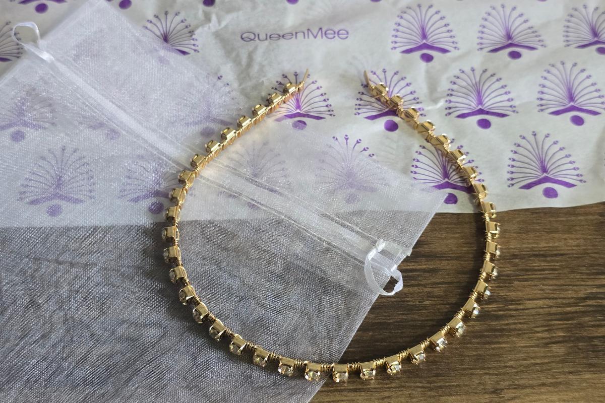 A gold headband with square embellishments lies on a wooden surface next to a sheer white drawstring pouch. Perfect for Christmas Gifts for Her, it’s beautifully presented with QueenMee tissue—ideal for anyone seeking festive Gift Ideas for Her.