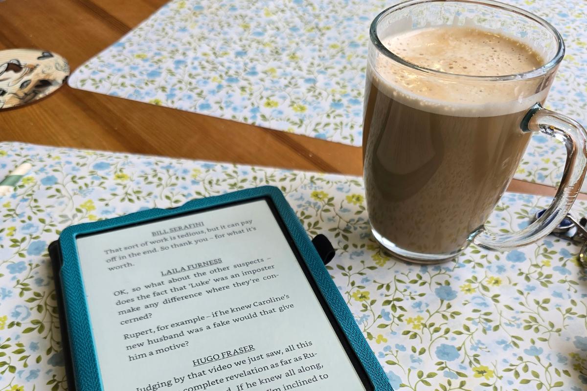 A tablet displaying an e-book lies on a floral-patterned tablecloth next to a clear glass mug filled with frothy coffee—perfect for those who love reading or enjoy reading in coffee shops. A coaster and part of a placemat are also visible in the background.