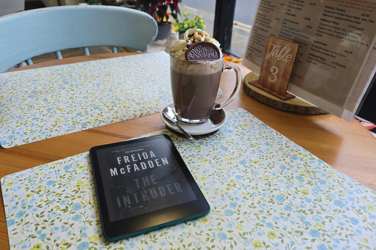 A tablet—an ideal pick for tech gifts—displays the cover of "The Intruder" by Freida McFadden on a floral placemat beside hot chocolate topped with whipped cream and a chocolate slice at a café table set for two.