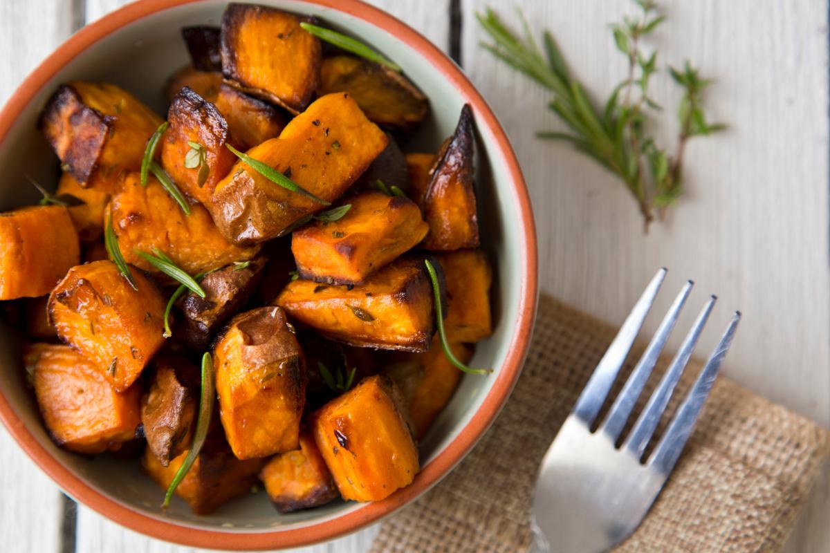 A bowl of perfect roasted sweet potatoes garnished with fresh rosemary sits on a light wooden table next to a fork resting on a burlap napkin.