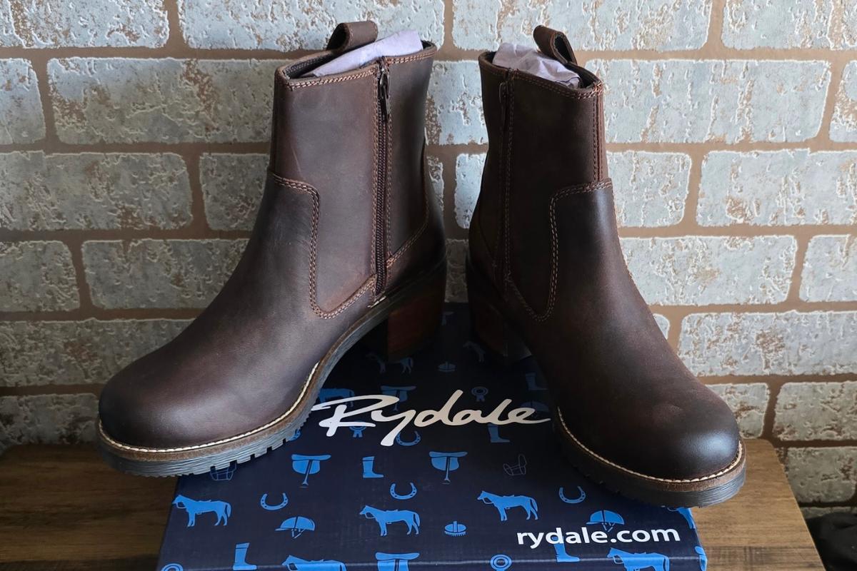 A pair of brown leather ankle boots with side zippers sits on a blue Rydale-branded box, making the perfect addition to your Ultimate Christmas Gifts list, set against a charming brick wall background.