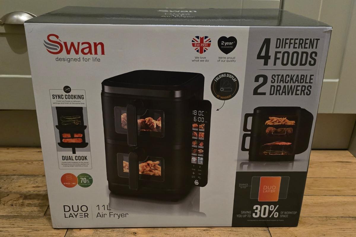 A Swan Duo Layer 11L Air Fryer box on a wooden floor, featuring 2 stackable drawers, sync cooking, dual cook, and energy saving—an ideal pick for any Kitchen Gifts list. The packaging shows the air fryer and delicious food inside.