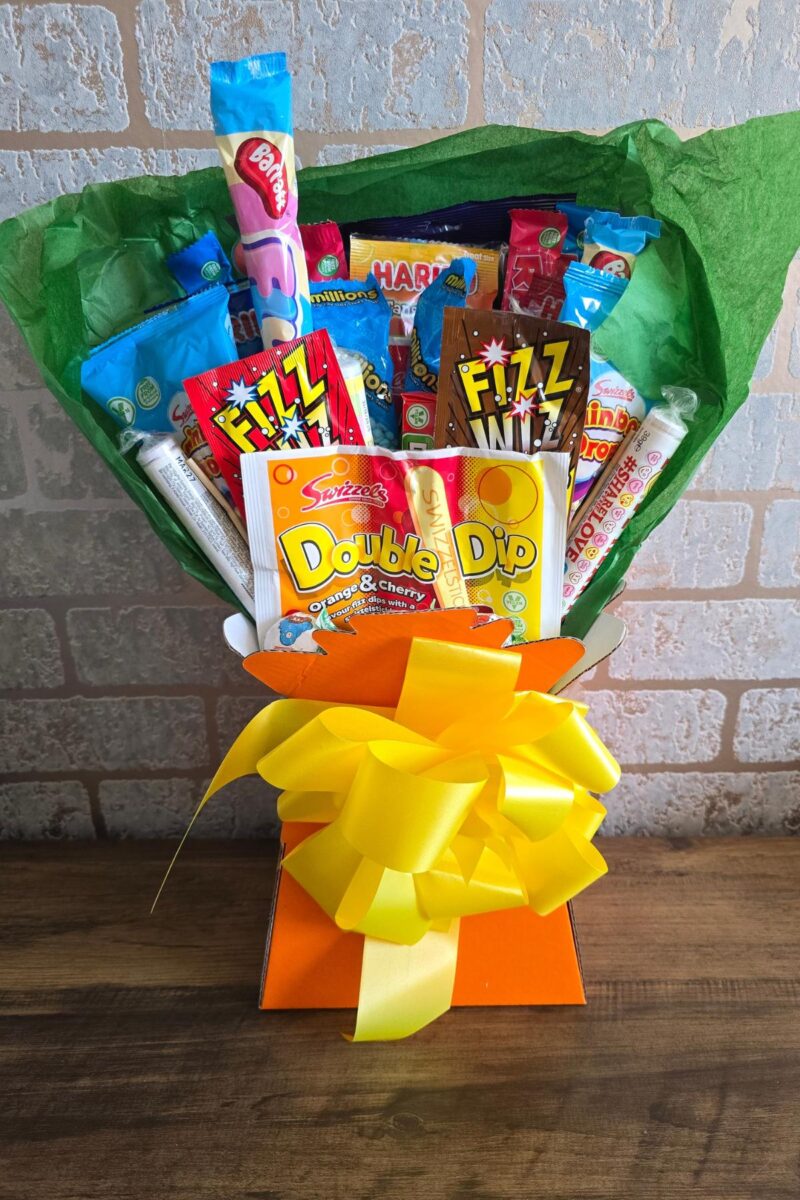 A vibrant candy bouquet features assorted colorful sweets and chocolate bars arranged in green tissue paper with a large yellow bow, making it a perfect choice for family gifts and holiday presents, displayed against a light brick wall.