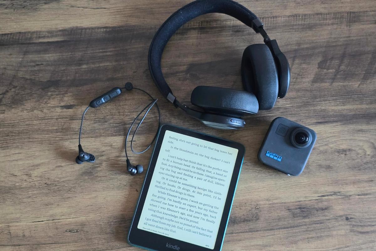 A Kindle e-reader displaying text, a pair of wireless headphones, wireless earbuds, and a GoPro camera rest on a wooden surface—perfect tech gifts for any gadget lover.