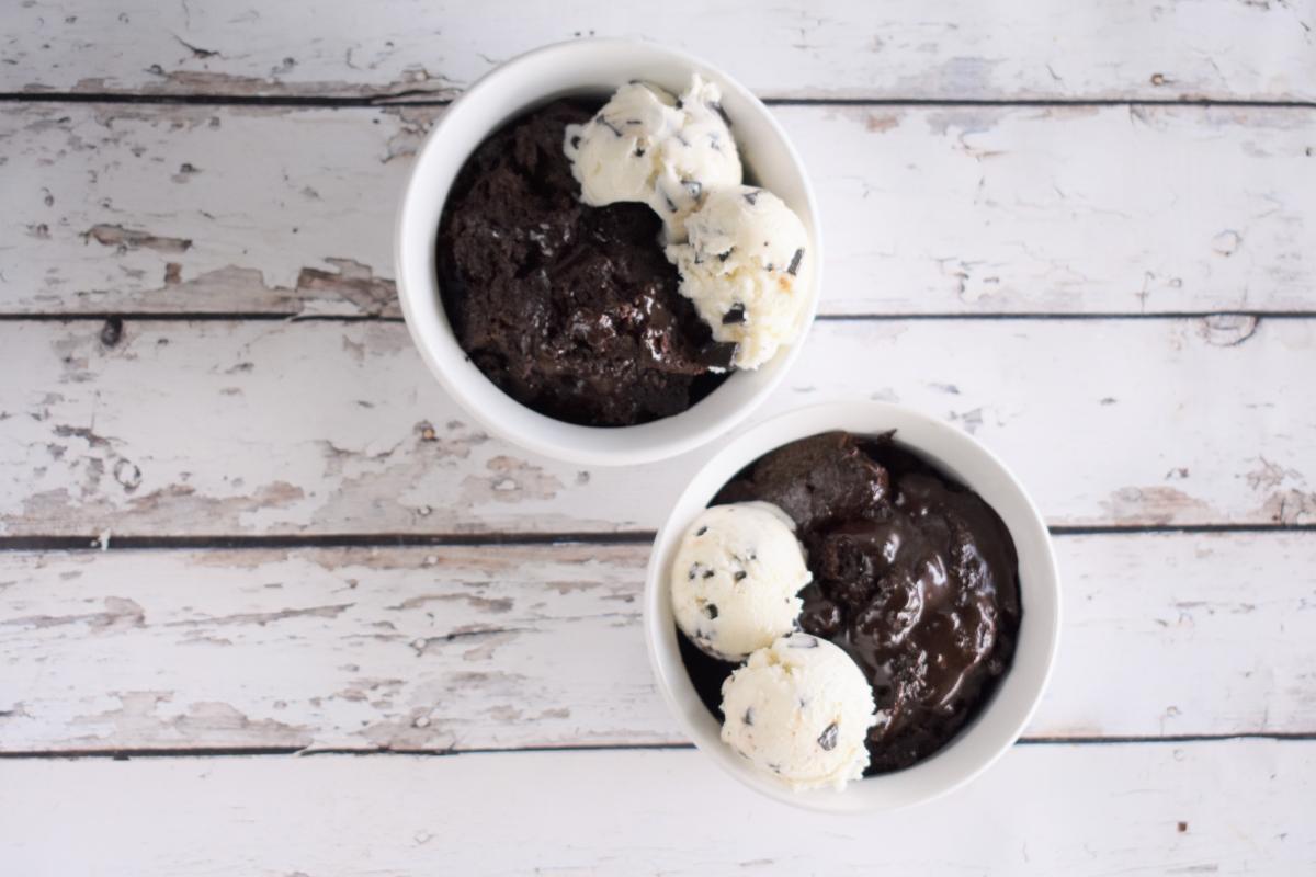 Two white bowls filled with slow cooker chocolate cake, topped with scoops of vanilla ice cream and chocolate chips, placed on a rustic white wooden surface.