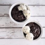 Two white bowls sit on a rustic white wooden surface, each filled with rich slow cooker chocolate cake and topped with two scoops of cookies and cream ice cream.