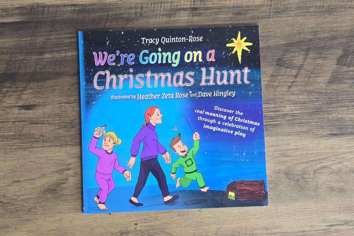 A children's book titled "We're Going on a Christmas Hunt" lies on a wooden surface. Perfect for kids holiday gifts, its cover shows three children holding hands under a star, with one in a green onesie and a treasure chest nearby.