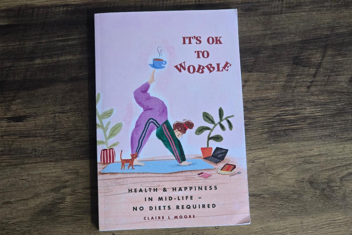 A book titled "It's OK to Wobble" features a woman doing yoga with a teacup on her foot, cat nearby, and plants, perfect for any Christmas Gift Guide. The subtitle reads "Health & Happiness in Mid-Life – No Diets Required.