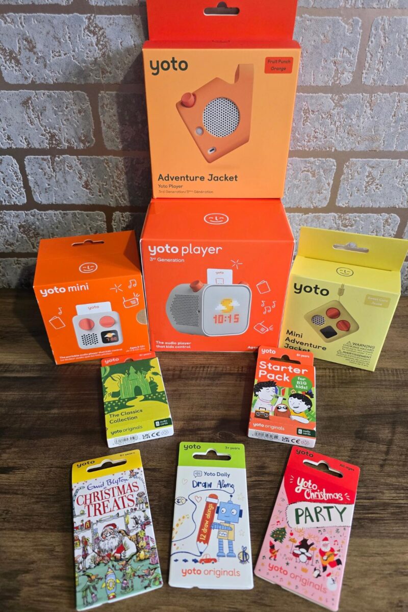 A selection of Yoto audio player products and accessories on a wooden surface, perfect for any children's gift guide. Includes a Yoto Player, Yoto Mini, Adventure Jacket case, and colorful audio cards. A brick wall is in the background.