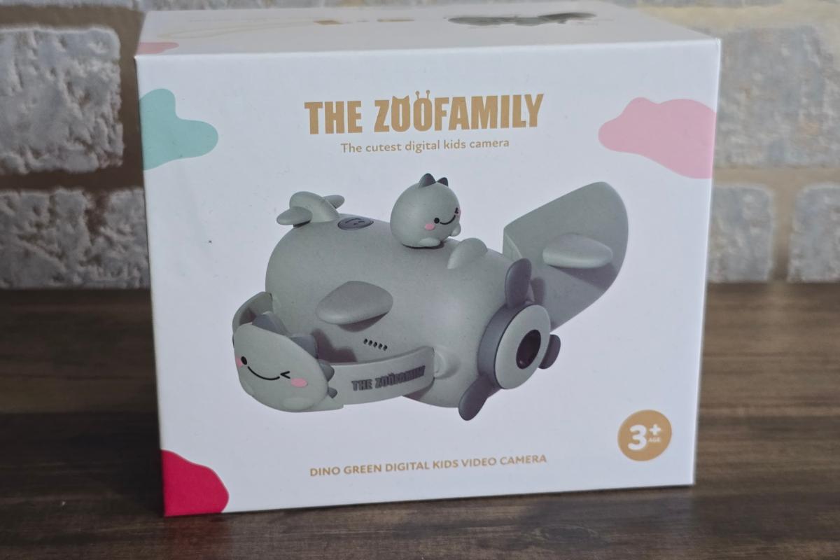 A box for "The Zoofamily" Dino Green Digital Kids Video Camera, decorated with cartoon animal and plane designs, sits on a wooden surface against a brick wall—an ideal pick for any children's Christmas gifts list.