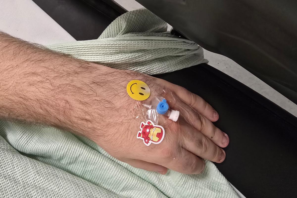 A hand with a cannula inserted, covered in transparent tape decorated with a smiley face sticker and an Iron Man sticker, rests on a light green hospital blanket—an image reflecting thoughtful hospital support for adults with learning difficulties.