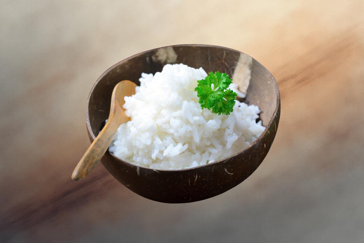 A wooden bowl filled with homemade coconut rice, garnished with a sprig of parsley and accompanied by a small wooden spoon, against a blurred background.