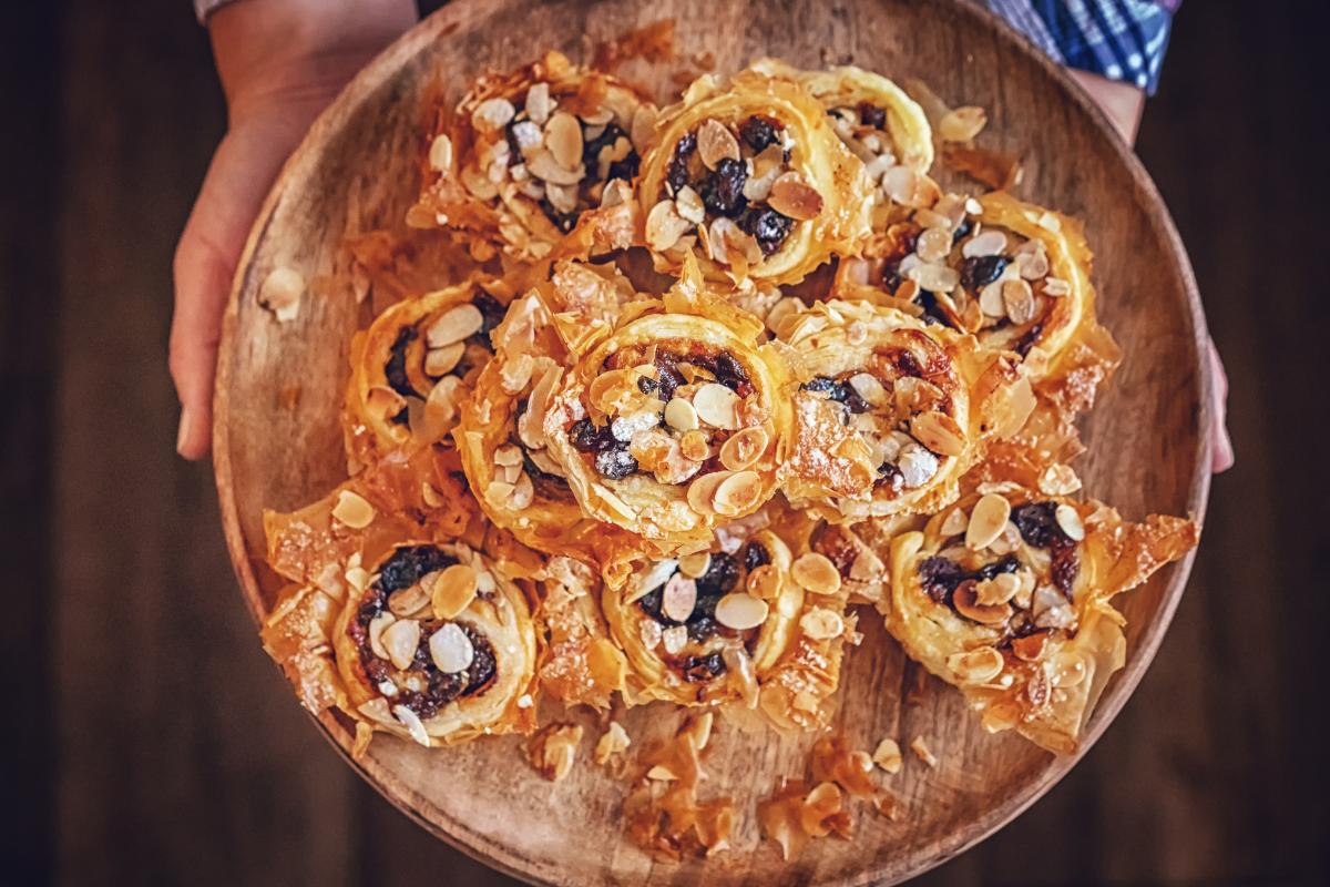A person holds a wooden platter topped with several round mince pies, each garnished with sliced almonds and sultanas, on a rustic background.