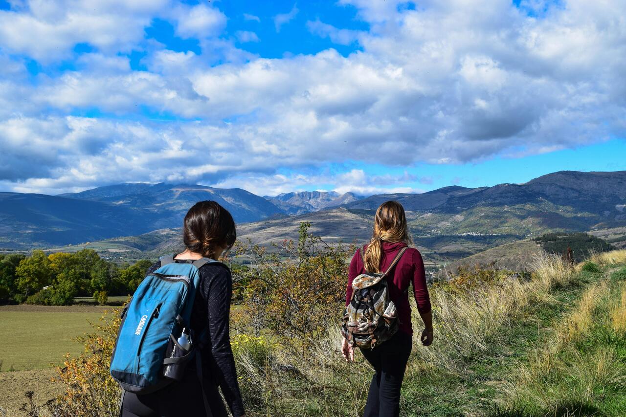 Two women with rucksacks hike along a grassy trail overlooking rolling hills and distant mountains, enjoying the mind and body benefits of long-distance walking under a partly cloudy blue sky.