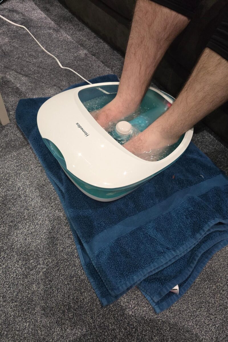 A person soaks their feet in a heated Homedics Foot Spa filled with water, placed on a blue towel atop a carpeted floor. The spa is plugged in and bubbles are visible around their feet, perfect for any foot spa review.