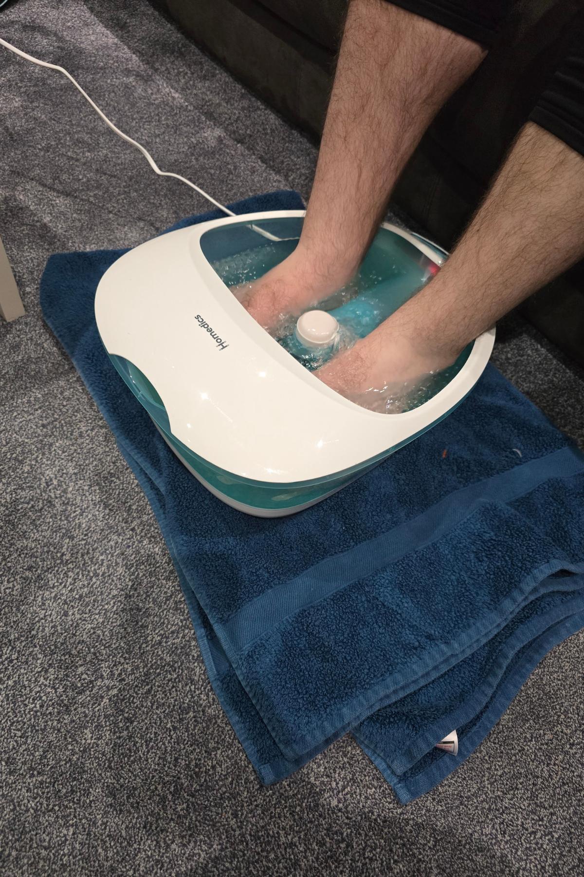 A person soaks their feet in a heated Homedics Foot Spa filled with water, placed on a blue towel atop a carpeted floor. The spa is plugged in and bubbles are visible around their feet, perfect for any foot spa review.