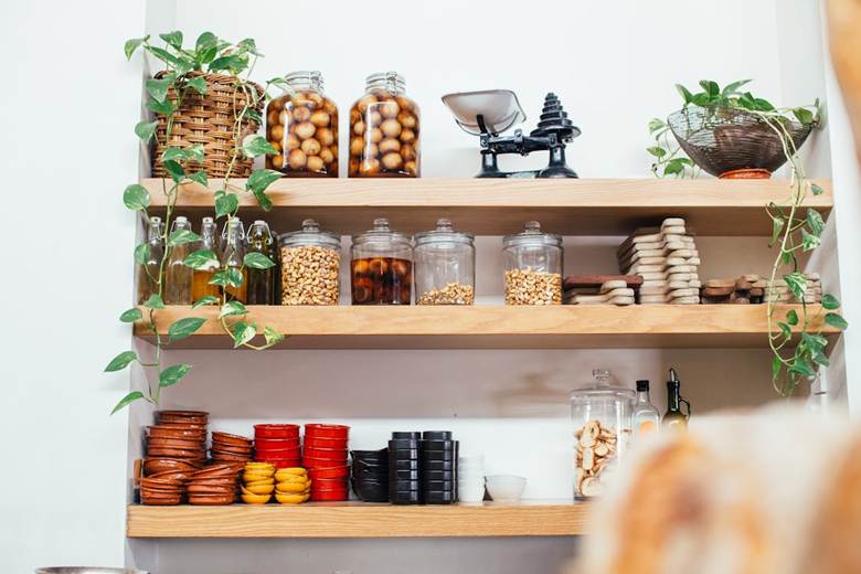 Wooden shelves with various jars of dried goods, bottles, a black vintage scales, stacked bowls, and potted trailing plants, all arranged neatly in a bright kitchen setting.