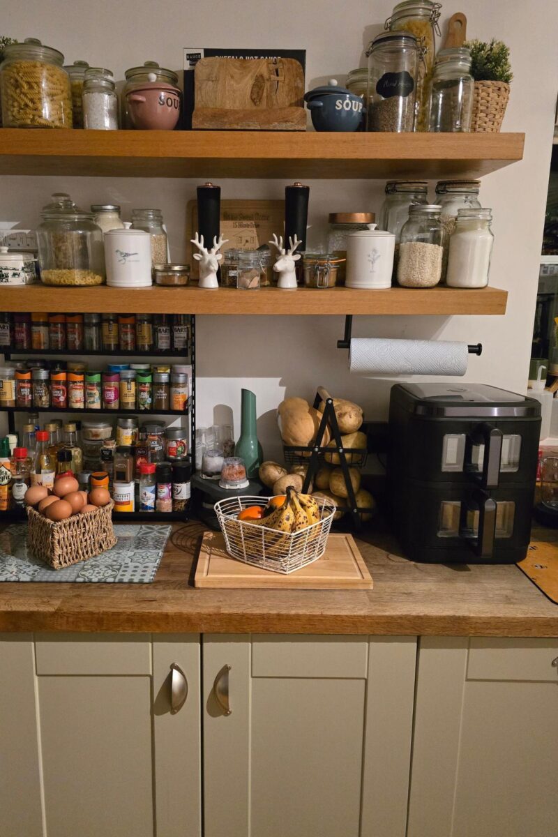 A cosy kitchen worktop with baskets of eggs and fruit, spices on a rack, jars on shelves, kitchen roll, and a sleek black Swan 11L Duo Layer Air Fryer. The shelves showcase grains, pasta, and charming kitchen decor.