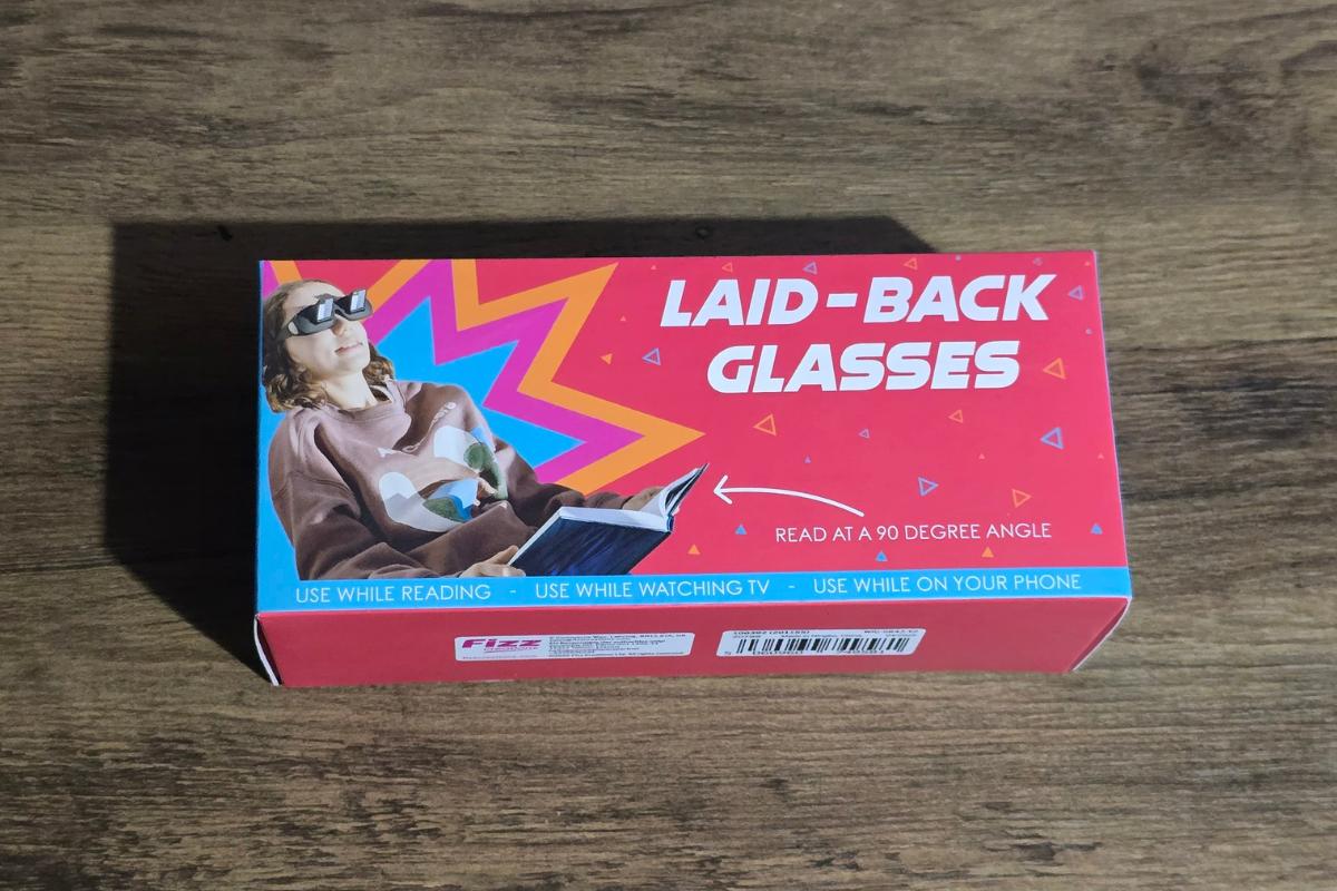A red box labelled "Laid-Back Glasses" from Wicked Uncle sits on a wooden surface, showing a person using prism glasses to read and relax. Perfect to win comfort whilst reading, watching TV, or using your phone whilst lying down.