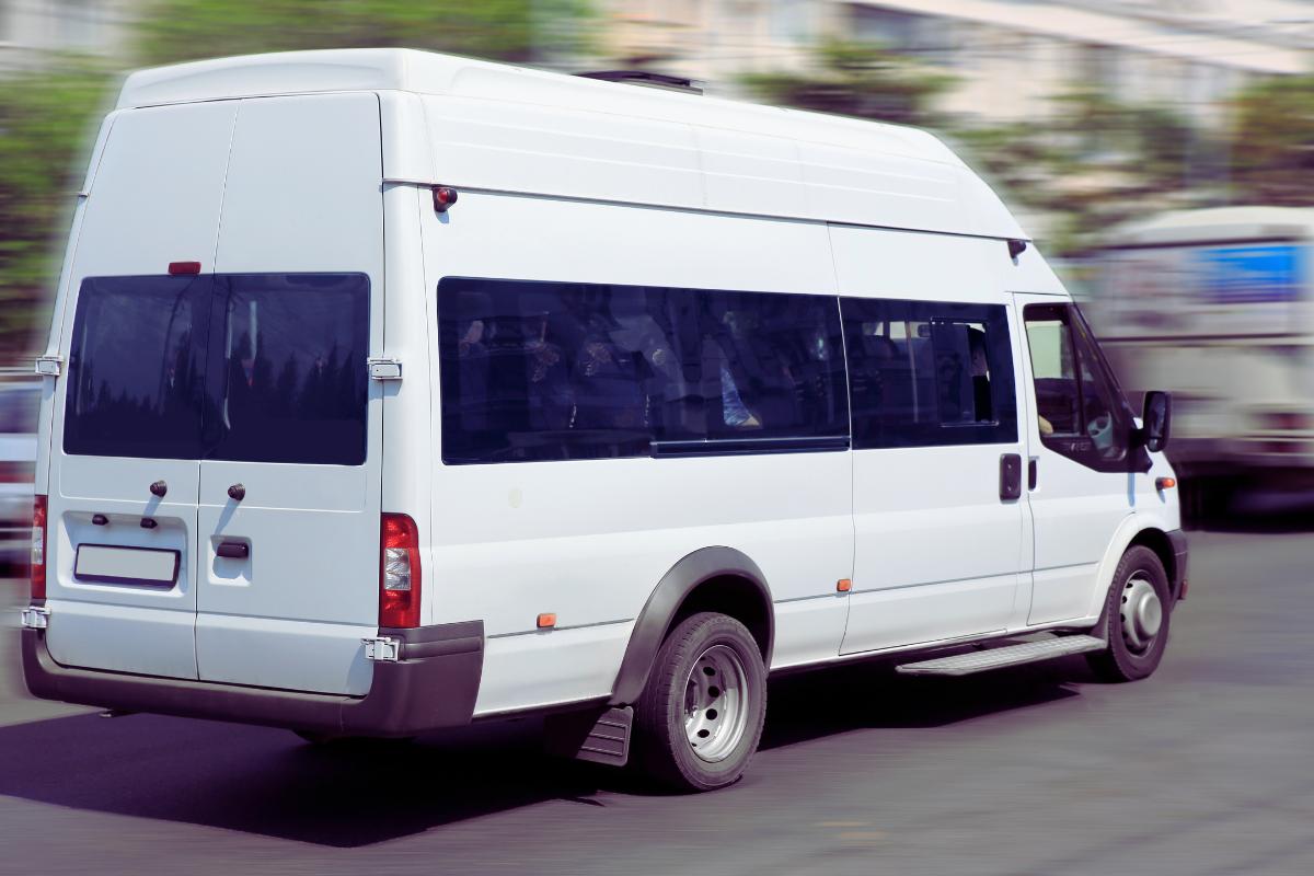 A white passenger minibus with tinted windows is driving on a city street, surrounded by blurred buildings and vehicles, capturing the essence of Organising Group Transport in Bradford.
