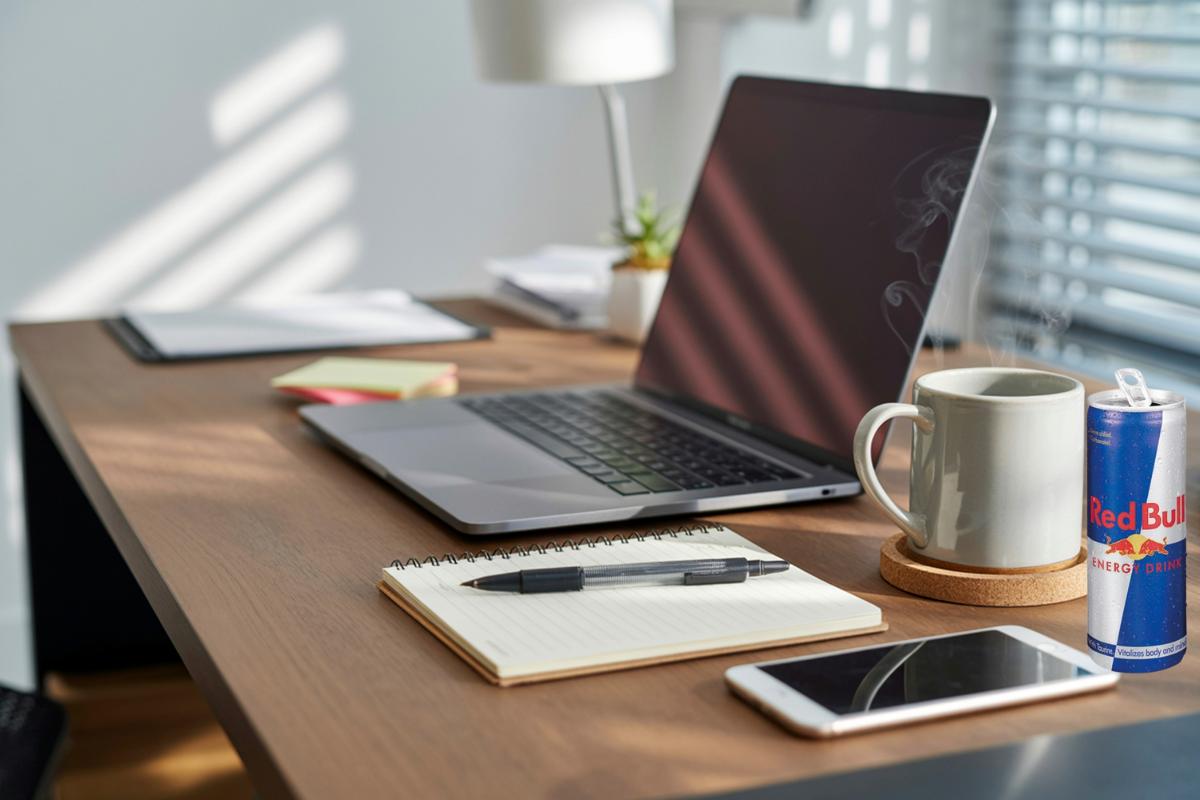 A tidy desk with a laptop, notepad and pen, mobile phone, closed Red Bull can, steaming mug, and documents. Sunlight streams through window blinds—enjoy a bright workspace atmosphere and consider healthy alternatives for your energy boost.