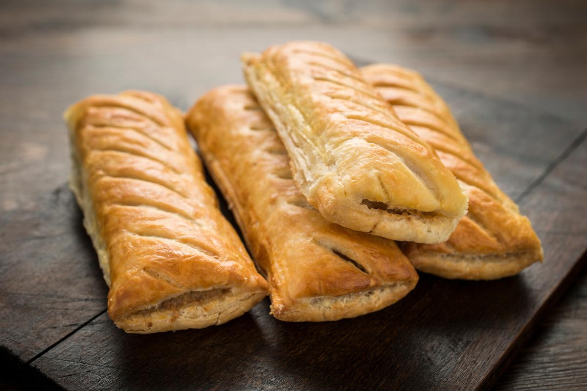 Four golden-brown sausage rolls are arranged on a dark wooden surface, showing flaky layers and slits on top—an enticing treat, though perhaps not the best choice if you’re watching your health.