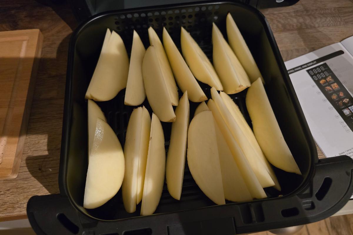 Sliced potato wedges arranged in a single layer inside a Swan 11L Duo Layer Air Fryer basket, ready to be cooked. The air fryer sits on a wooden surface with a chopping board and paper nearby.