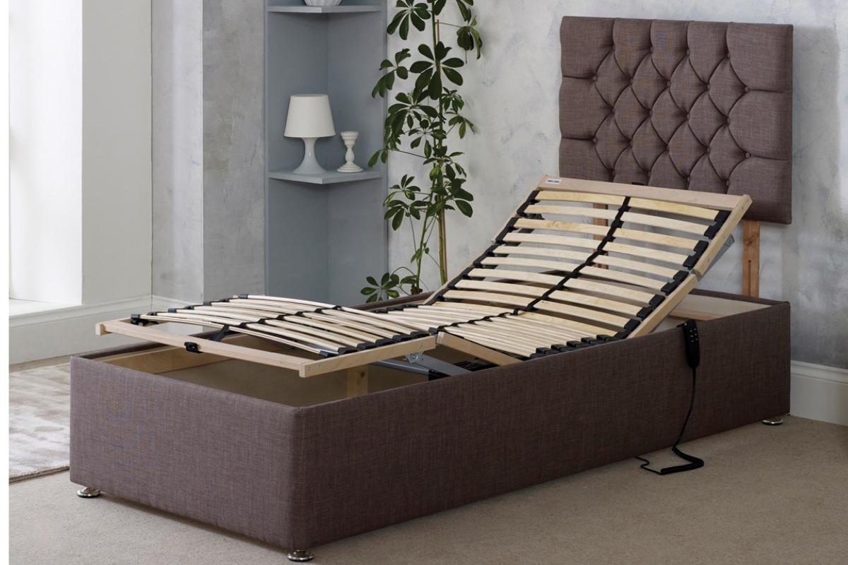 A single adjustable bed with a brown upholstered frame and headboard is shown with the head and foot sections raised—an ideal choice for accessible bedroom design. A remote control is attached to the bed, with a plant and small shelf in the background.