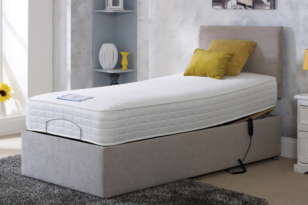 A single adjustable bed with a thick white mattress, light grey upholstered base, beige headboard, and yellow accent cushions creates an accessible bedroom. Light decor and soft natural light enhance the inviting home design.