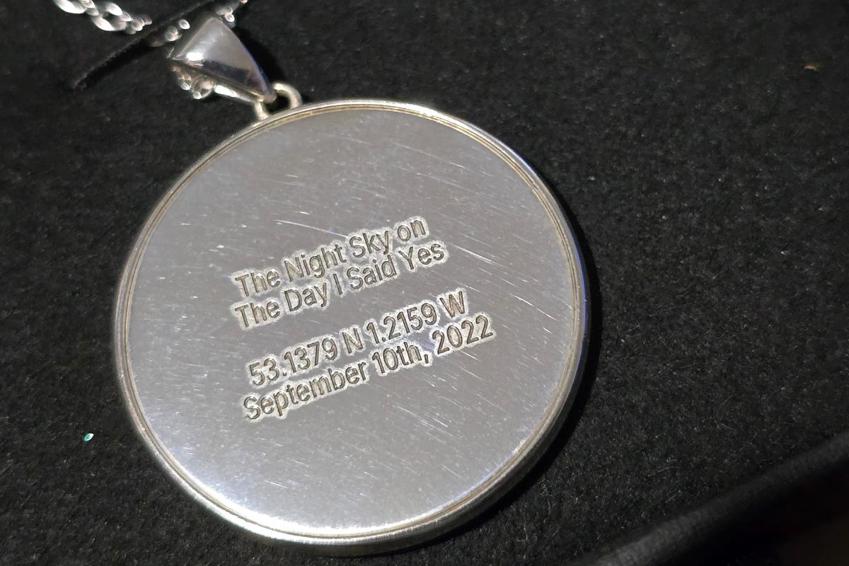 A round silver night sky necklace pendant inscribed with "The Night Sky on the Day I Said Yes 53.1379 N 124.159 W 10th September 2022," resting elegantly on a black surface.