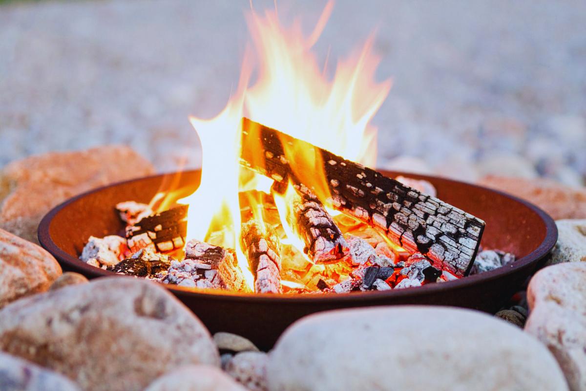 A close-up of a campfire burning in a metal fire pit, with glowing orange flames and charred logs, surrounded by large stones on a pebbled surface outdoors—an ideal way to choose firepit set-ups like clever firepit options in the UK.