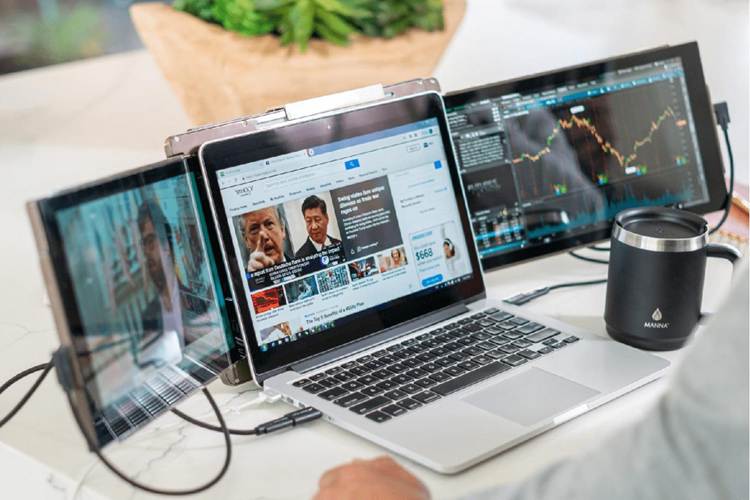 A laptop with two portable monitors attached on either side displays news and share charts. A person’s hand rests nearby, and a black insulated mug sits on the desk next to the laptop.