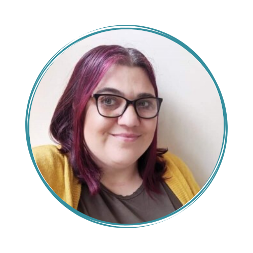 A person with shoulder-length, dark hair with purple highlights, wearing glasses, a brown top, and a yellow cardigan, smiling at the camera. The photo is framed by a teal circular border.