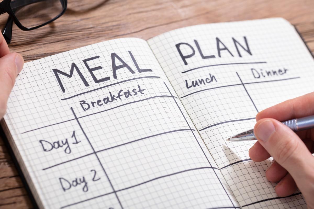 A person writes a 7-day meal plan in a squared notebook labelled "Meal Plan," with sections for breakfast, lunch, and dinner across multiple days. Glasses and a pen rest on the wooden surface.