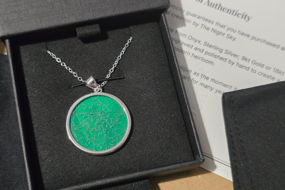 A silver night sky necklace with a round green pendant featuring a constellation map design is displayed in an open black jewellery box, with a certificate of authenticity partially visible in the background.