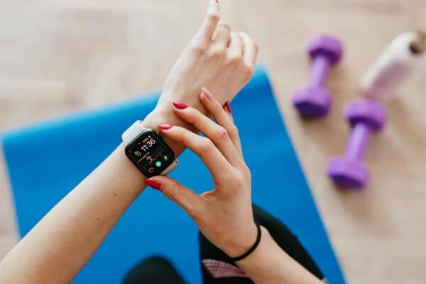 Person checking a smartwatch with fitness stats while sitting on a blue exercise mat, with purple dumbbells and a water bottle nearby.