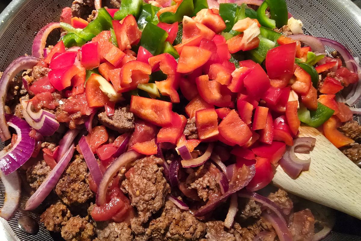 Chopped red and green peppers, sliced red onions, and cooked minced beef are mixed together in a metal colander for a healthy meal. A wooden spoon is partially visible on the right side of the image.