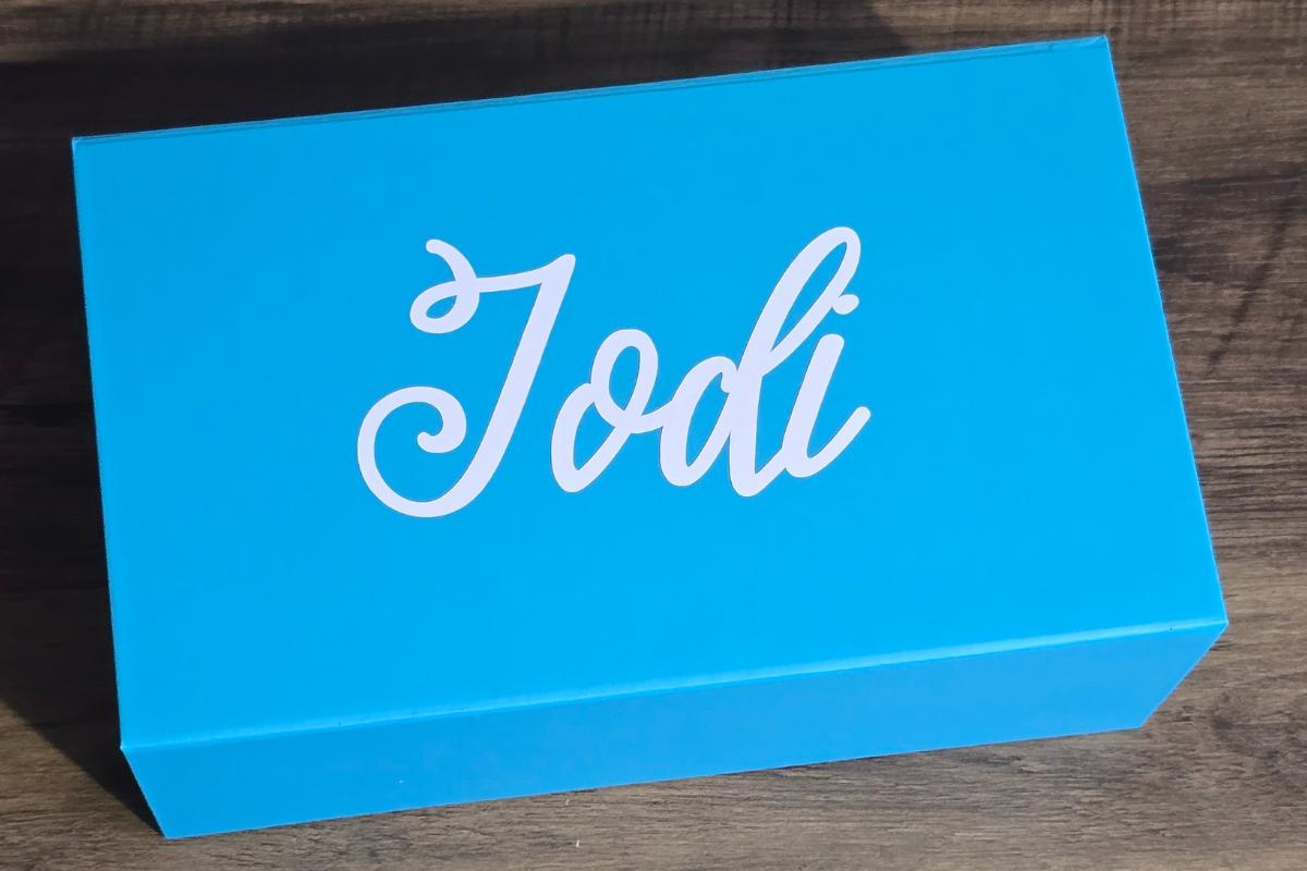 A bright blue rectangular box from Something Blue Ladies with the name "Jodi" in white cursive letters on the lid, sitting on a wooden surface. Perfect for bridesmaids or as part of your How I Asked moment.