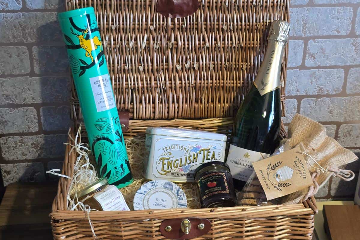 A wicker hamper basket filled with gourmet items—perfect for time together. Includes a teal tube with a stag design, english tea tin, sparkling wine, jam jars, hessian bag, and straw packing, all set against a charming brick wall.