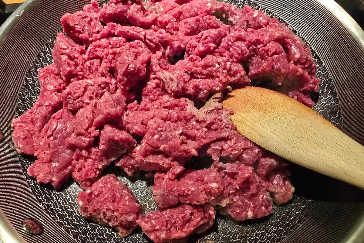 Raw minced beef in a frying pan, with a wooden spatula resting on the meat, ready to be cooked—perfect for pairing with spicy potato wedges for a delicious, healthy meal.