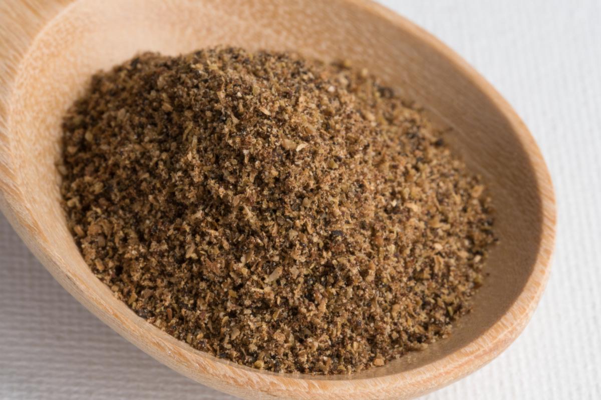 A close-up of a wooden spoon filled with ground black pepper, a classic warming spice often used in family meals, placed on a white textured surface. The pepper appears coarse and dark brown in colour.