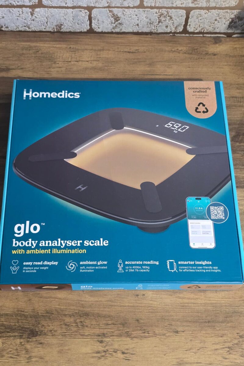 A boxed HoMedics glo Body Analyser Scale with ambient illumination sits on a wooden surface. The teal box highlights features like easy-read display, ambient glow, and smartphone connectivity—perfect for those wondering if it's worth the hype.