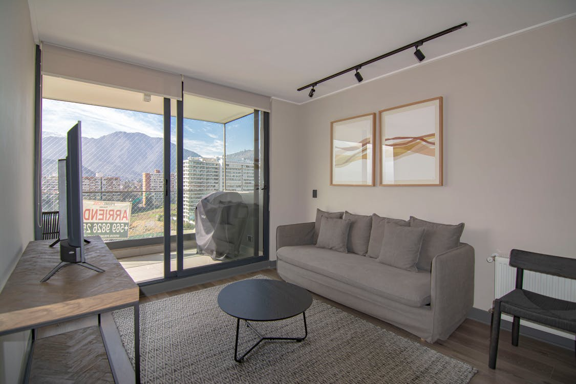 Modern living room with a grey sofa, round black coffee table, TV on a wooden stand, large window with city and mountain view, and framed abstract art on the wall.