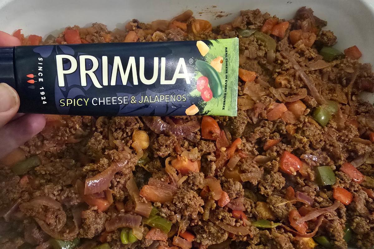 A hand holds a tube of Primula Spicy Cheese & Jalapenos spread above a pan of cooked minced beef with chopped onions and peppers—perfect for topping Cajun loaded potato wedges or creating a healthy meal bursting with flavour.