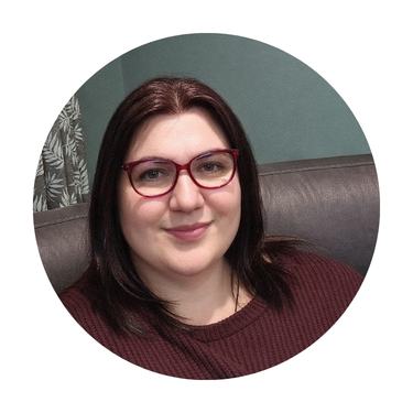 A person with straight, dark brown hair and red glasses smiles whilst sitting on a dark sofa. Showcasing a cosy lifestyle, they wear a maroon jumper against a teal wall with a leafy pattern partially visible.