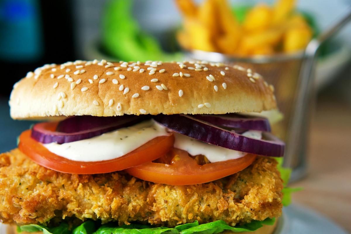 A close-up of a crispy chicken burger with lettuce, tomato slices, red onion rings, and creamy sauce in a sesame seed bun—perfect for a KFC Fakeaway night at home. Blurred chips in a metal basket are visible in the background.