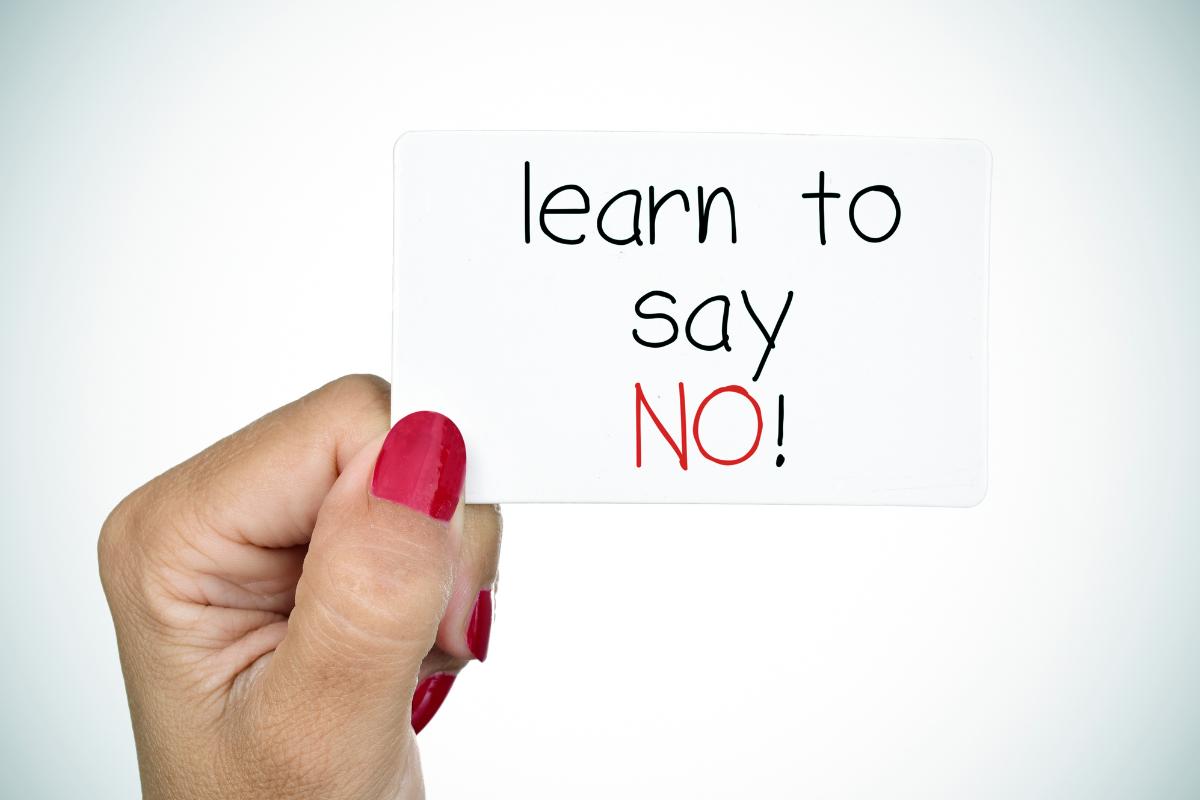 A hand with red-painted nails holds a white card that reads, in a full sentence, "learn to say NO!"—reminding you why it is OK to say no boldly when needed.
