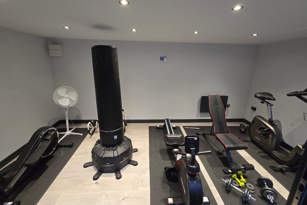 A home gym room with various exercise equipment, including a punch bag, weights, bench, Yosuda RM4 rowing machine, fan, and exercise bike, all arranged on wood and black flooring under ceiling spotlights.