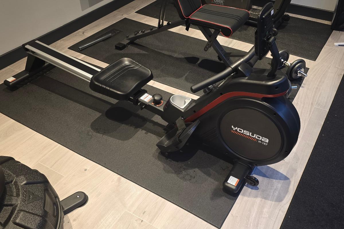 A black Yosuda RM4 rowing machine sits on a mat in a home gym with light wood floors. A bench and tyre are also visible nearby.